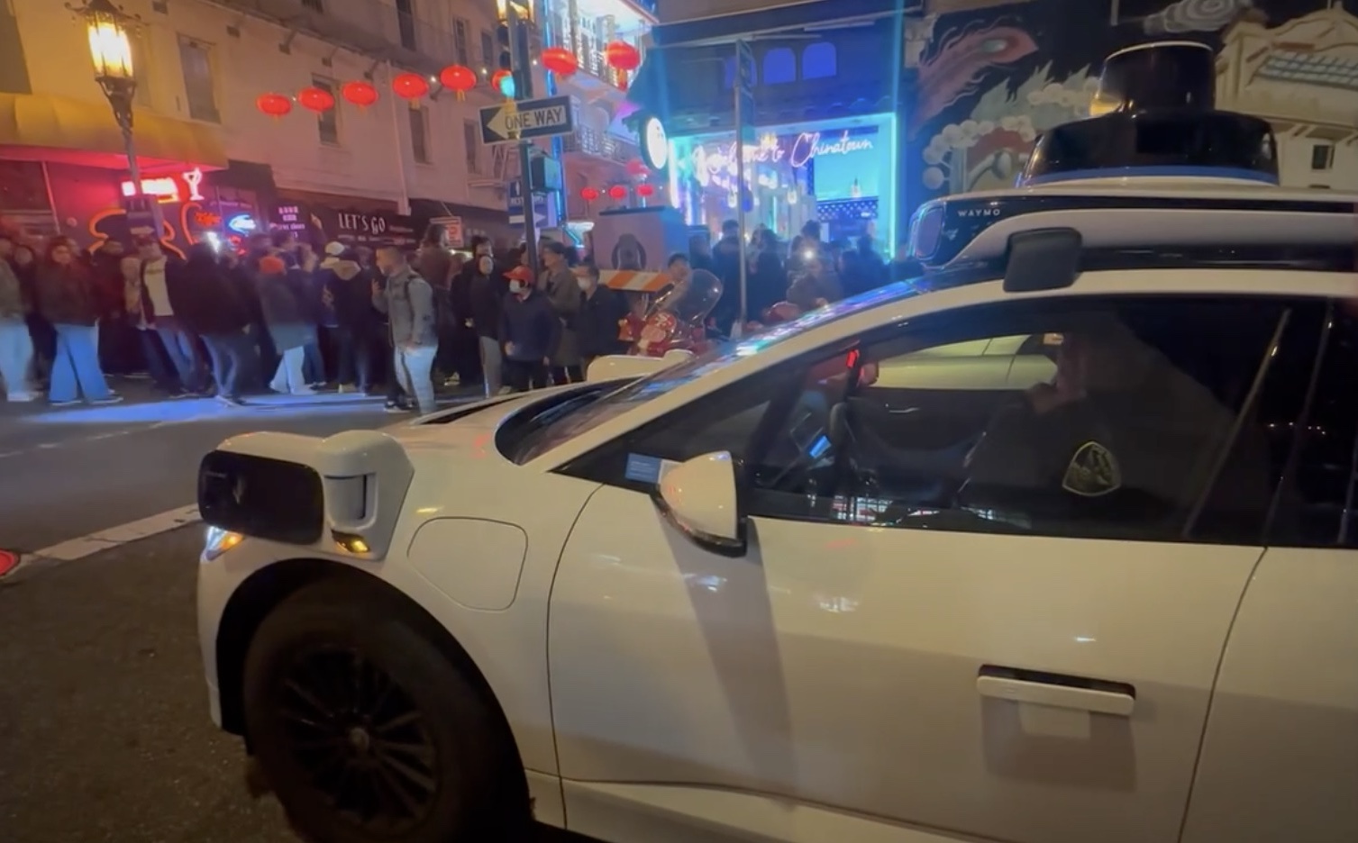 A sleek, autonomous Waymo white car paused at a bustling Chinatown intersection under the night sky, with people strolling and vibrant signs illuminating the background.